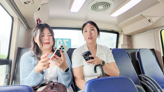 In a springtime Seoul bus, two Korean women in their twenties in casual attire sit side by side, laughing and chatting as city scenes pass by outside the windows. Seoul, Jongno District, spring. - Powered by Adobe