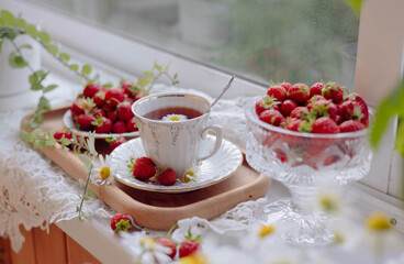 A cozy tea setting features a porcelain cup filled with tea next to a beautifully arranged bowl of fresh strawberries. The natural light enhances the inviting atmosphere near the window