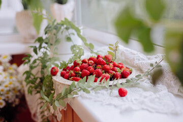 A decorative plate overflowing with fresh strawberries sits on a windowsill adorned with greenery and lace. The sunlight gently illuminates the vibrant red berries