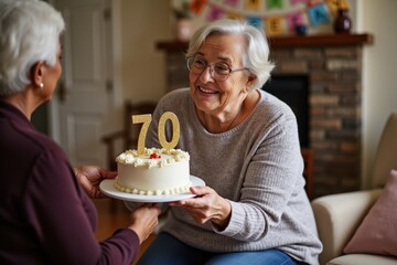 An old black woman is presenting a birthday cake with the number 70 to an old woman. They are in a cozy living room with a fireplace and birthday banners