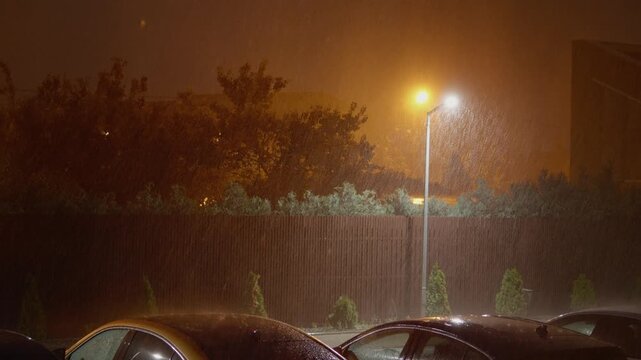 Intense storm with heavy rain and strong winds affects homes and parked cars. Dark clouds loom above while lightning strikes illuminate the night sky.