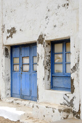 Fototapeta premium A faded blue door and matching window stand on a crumbling whitewashed wall. The aged textures tell a quiet story of time, abandonment, and Mediterranean charm.