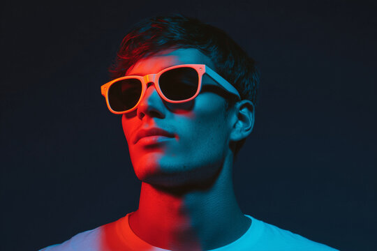 Guy standing in darkness wearing glowing neon sunglasses, illuminated with vibrant electric colors, creating a modern, stylish, edgy vibe with strong contrast in an urban, futuristic atmosphere - Powered by Adobe