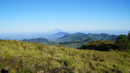 Fototapeta premium Wide view of green mountain landscape under clear blue sky, with distant volcano visible on the horizon.