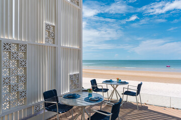 Stylish beachfront restaurant terrace with modern white block wall, marble tables and blue chairs on sunny day, offering relaxing outdoor dining experience