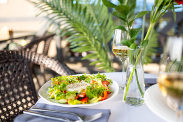 Fresh vegetable salad with cheese and white wine served on an outdoor table with wicker chairs and greenery, representing summer dining and relaxation.