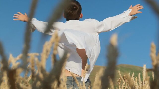Back view of a teenage boy with a fit body standing in golden ripe wheat at sunset, facing a green smooth volcano. His white unbuttoned shirt flutters in the wind, capturing a moment of freedom.