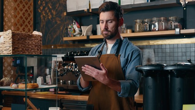 Glad Caucasian barista waiter male man guy bartender worker typing tablet order online app device tech in restaurant catering delivery employee looking at camera small business owner smiling cafeteria