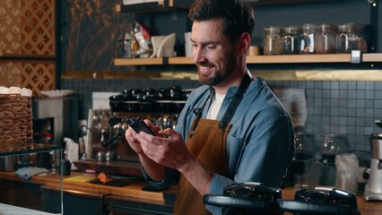 Happy Caucasian barista worker waiter guy scroll screen mobile phone employee male laughing in cafeteria bartender man typing smartphone cyberspace online small business Internet shopping social media