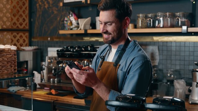 Happy Caucasian barista worker waiter guy scroll screen mobile phone employee male laughing in cafeteria bartender man typing smartphone cyberspace online small business Internet shopping social media