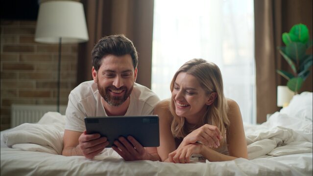 Laughing Caucasian family married couple holding tablet in bedroom love cheerful boyfriend girlfriend typing device woman purchasing goods app Internet man remote shopping together morning cyberspace