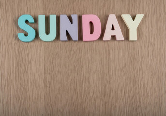 Fototapeta premium Colorful wooden letters spelling out the word SUNDAY are arranged in a horizontal line across a light brown wooden surface