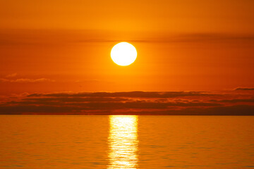 Panorama of a bright orange sunset with the sun fully visible over the sea and long clouds on the horizon