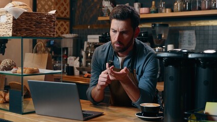 Pensive Caucasian entrepreneur barista employee male browsing Internet app laptop commerce small business owner thinking in cafeteria manager choosing guy online shopping cafe man waiter typing gadget