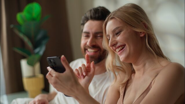 Happy Caucasian couple family using mobile phone together in bedroom boyfriend girlfriend laughing pair scroll smartphone woman man purchasing goods online shopping Internet browse web apps connection