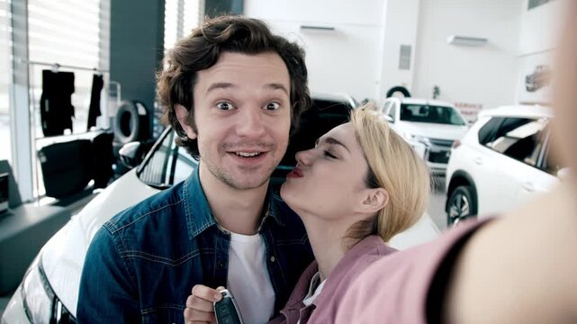 Happy couple taking a selfie in a car dealership, showcasing excitement after purchasing a new vehicle with cars in the background
