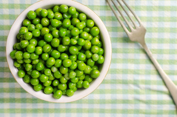 Boiled green peas in a ceramic bowl with a fork on a checkered tablecloth. Natural food concept. Vegetarian and vegan food. Horizontal orientation. Selective focus. Top view.