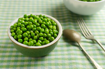Boiled green peas in a ceramic bowl with a fork and spoon on a checkered tablecloth. Natural food concept. Vegetarian and vegan food. Horizontal orientation. Selective focus.
