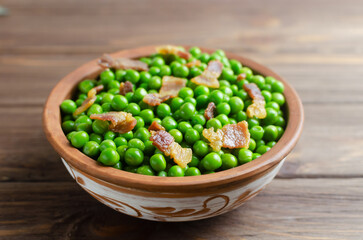 Boiled green peas with fried crispy bacon in a brown ceramic bowl on a wooden table. Old recipe concept. Simple dishes. Rustic style. Horizontal orientation. Selective focus.
