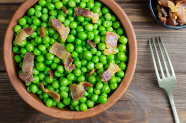 Boiled green peas with fried crispy bacon in a brown ceramic bowl with a fork on a wooden table. The concept of an old recipe. Rustic style. Horizontal orientation. Selective focus. Top view.