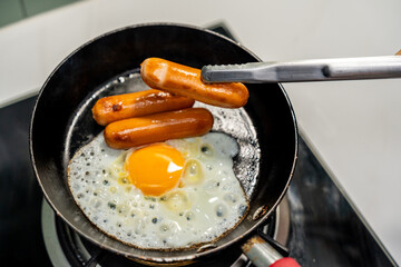A hand expertly flips sausages in a hot pan while perfectly fried eggs cook alongside them. Sausages sizzle and brown next to sunny-side-up eggs.