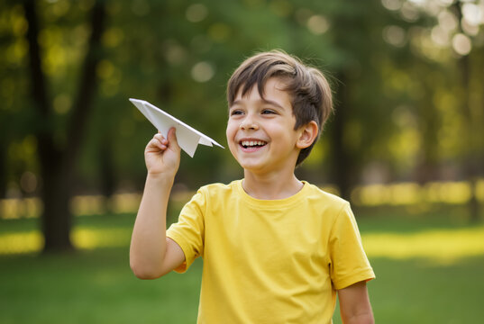 Happy boy playing with a paper airplane in the park. Laughing child in a yellow t-shirt launching a paper toy outdoors. Childhood, dream and imagination concept.