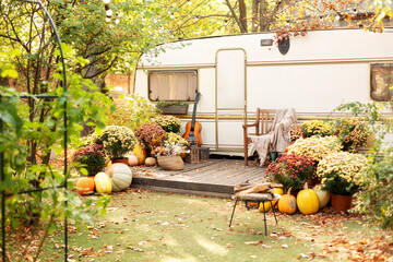 Interior cozy yard campsite with fall flowers potted and pumpkins decorated for Halloween. Armchair near caravan trailer. Exterior RV house porch with garden furniture. Campsite with firepit in garden