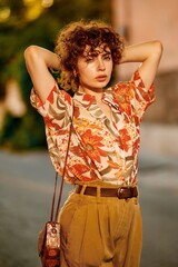 A woman with curly hair poses confidently in a floral shirt and stylish outfit