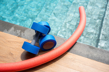 Two pieces of aquatic exercise equipment, a buoyant dumbbell and a swim noodle, are positioned by the edge of a swimming pool. Water-based fitness, hydrotherapy, and rehabilitation exercises.