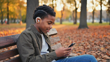 Teenage African American boy with short curly hair and hearing aid sitting on a wooden bench in autumn park, wearing khaki jacket and jeans, smiling at smartphone, warm golden sunlight and fallen leav