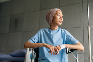 A cancer patient sits in a wheelchair with an IV drip looking stressed and sad. Asian Woman receiving IV therapy in a wheelchair showing visible signs of stress and depression