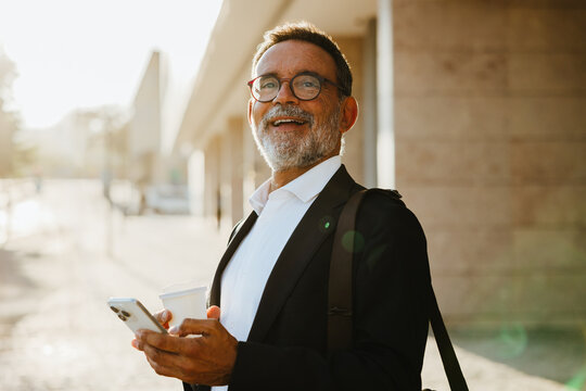 Senior businessman outdoors holding smartphone and coffee cup with a cheerful mood - Powered by Adobe