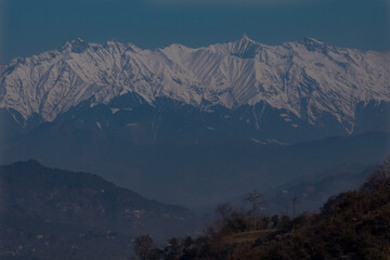 Snow covered mountain peaks with forest hills in Himalayas. A breathtaking view of snow-covered mountain peaks towering high above lush green forested hills, captured in Himachal Pradesh, India.