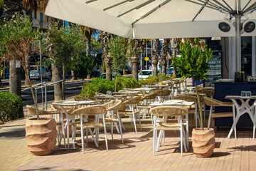 Cozy cafe with wicker furniture, white tables, and greenery under a canopy with fans on a sunny Mediterranean street lined with palm trees.