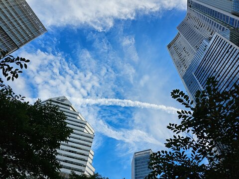 Modern skyscrapers framing a vibrant blue sky with scattered clouds and a dramatic white vapor trail, viewed from a low angle in an urban setting.