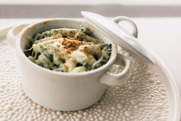 Delicious baked dish, topped with melted cheese and seasonings, served in a white ceramic pot with a lid. Baked green asparagus beans. Elegant presentation of the dish on a beautiful white plate.