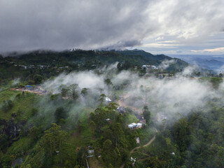 Aerial view of a misty landscape where clouds dance among the lush green hills and small buildings, creating a serene and ethereal scene, Ella, Sri Lanka.