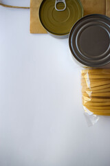 Top view of canned food with pull tab, brown packaging, and a transparent bag of pasta, arranged on a clean white surface, showcasing food preparation essentials