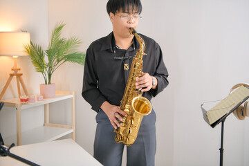 Fototapeta premium Young Asian man playing saxophone indoors in a cozy room with soft lighting