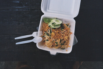 Pad thai noodles served in a takeaway box with plastic cutlery on a dark wooden table