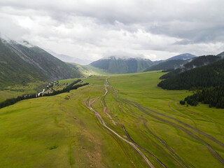 A wide aerial view of a green mountain valley with a few dirt trails winding through the landscape and cloudy peaks in the distance