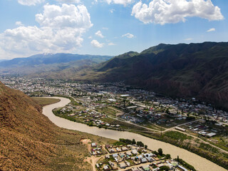 An aerial view of Naryn city and the winding Naryn river, surrounded by mountains under a bright, cloudy sky in Kyrgyzstan