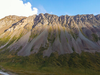 A wide drone shot of a steep, textured mountain ridge with rocky slopes and green grassy areas under a blue sky with some clouds