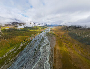 A wide aerial shot of a braided river flowing through a vast green mountain valley, with low clouds hugging the distant peaks