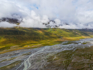 A wide aerial shot of a vast braided river flowing through a remote mountain valley with green and yellow grass under a dramatic cloudy sky