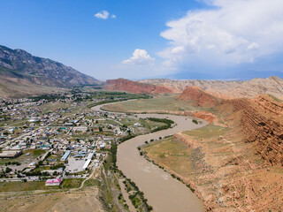 A wide aerial view of the Naryn river, with its winding path and a city on its bank, surrounded by mountains and red rocky hills