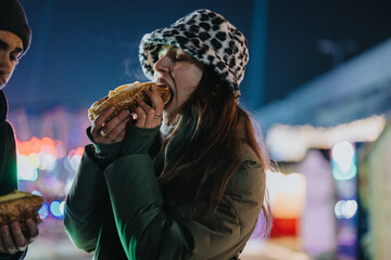 A woman in a cozy outfit eating a hotdog under colorful lights during winter.
