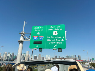 View from an open-top tour bus showing a Miami downtown traffic sign against a bright blue summer sky, with passengers' heads just visible in the foreground.