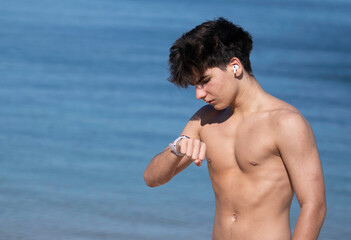 Portrait of shirtless young man wearing earbuds and checking the time on his smartwatch at the beach on workout time with copy space.