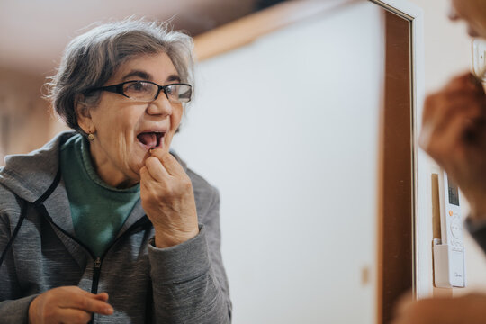 Elderly woman engaged in flossing her teeth while looking in a mirror, emphasizing self-care and healthy oral hygiene practices. - Powered by Adobe